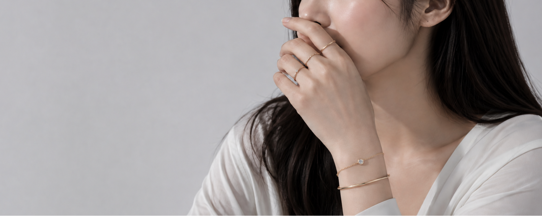 A woman in a white top wearing delicate gold jewelry, including a thin gold ring on her finger and a fine gold chain bracelet on her wrist, against a plain light gray background.