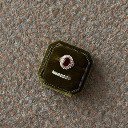 A silver ring set with a round red gemstone and a matching silver band, displayed inside an octagonal dark green velvet jewelry box on a textured gray carpet.